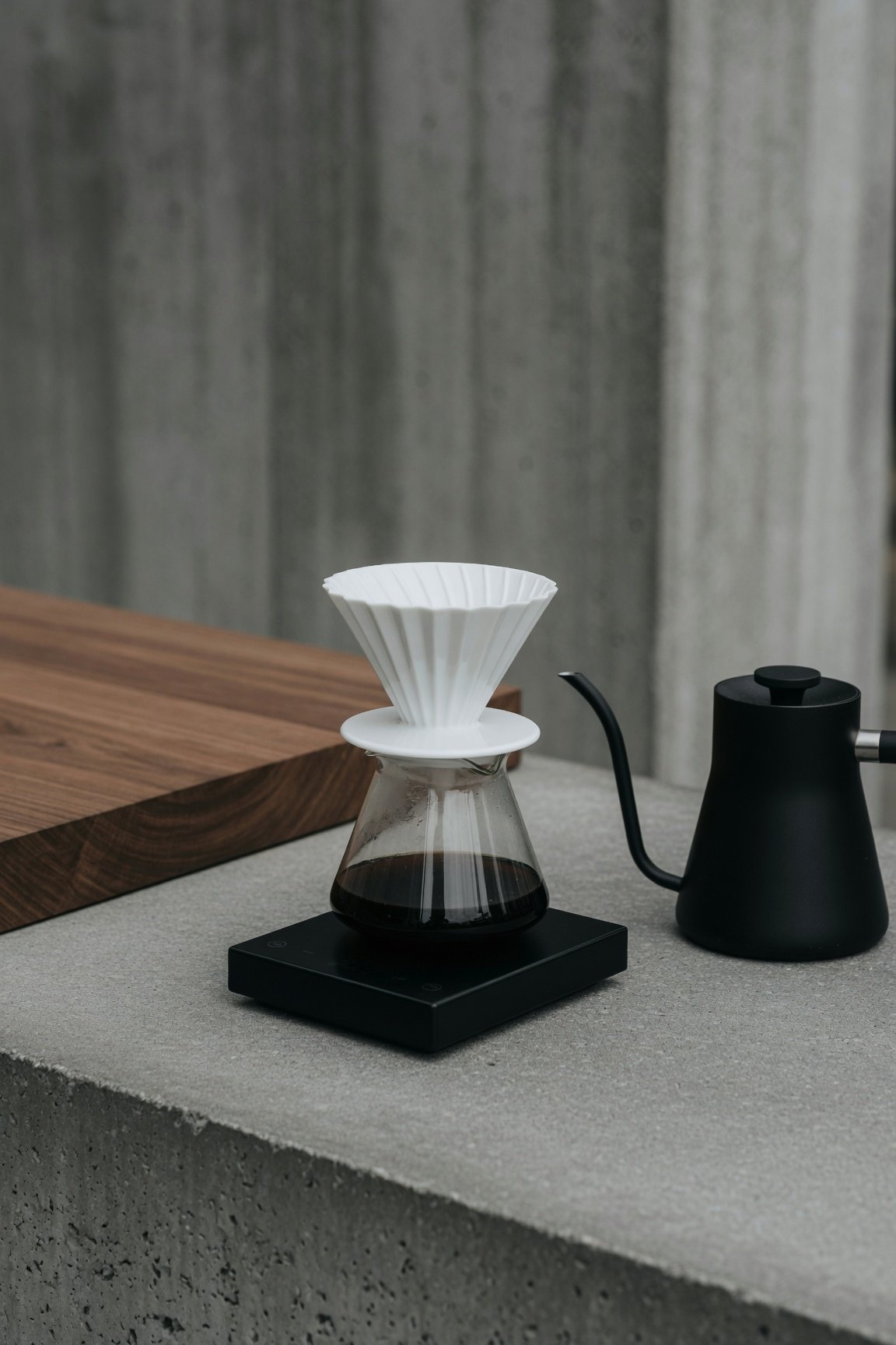 Dripper de cerámica blanca sobre balanza negra con jarra de vidrio y pava de cuello de cisne, en mesada de hormigón